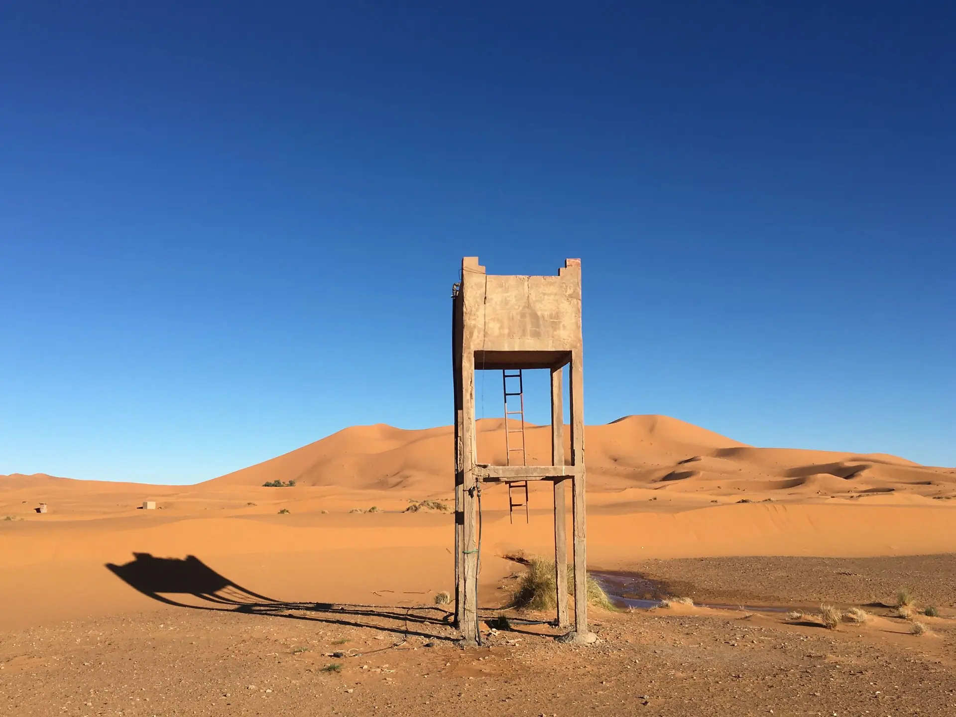 Concrete structure standing amidst the vast Merzouga sand dunes under a clear blue sky.